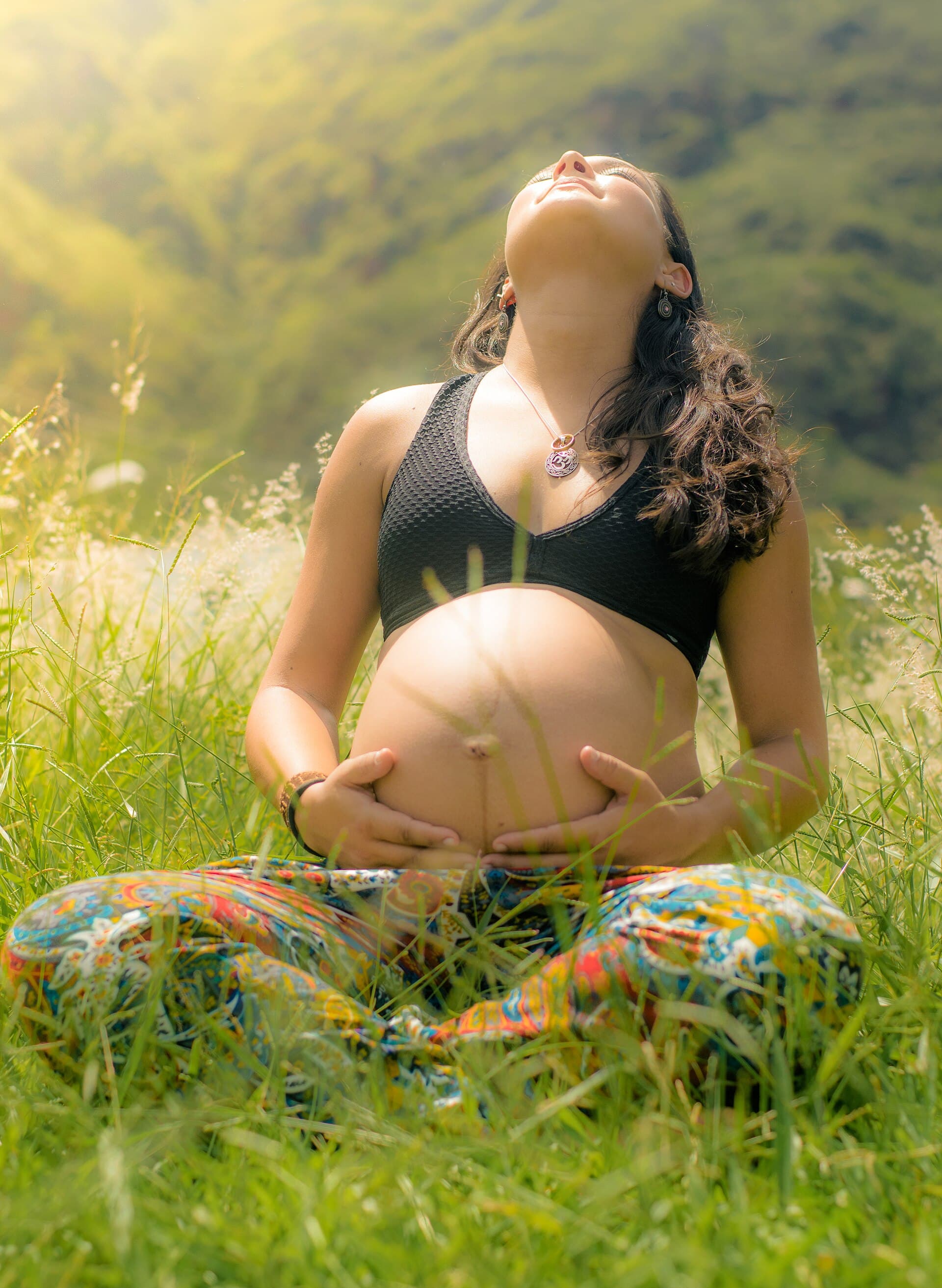 Gestante sentada na grama, de olhos fechados, com as mãos sobre a barriga